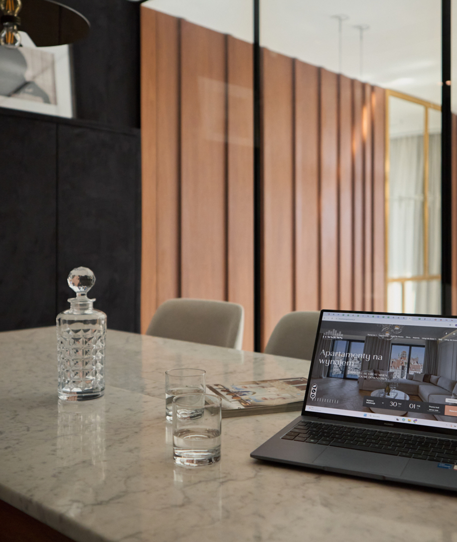 Tavolo in marmo in suite moderna, caraffa di cristallo, bicchieri e laptop con pagina di affitti.