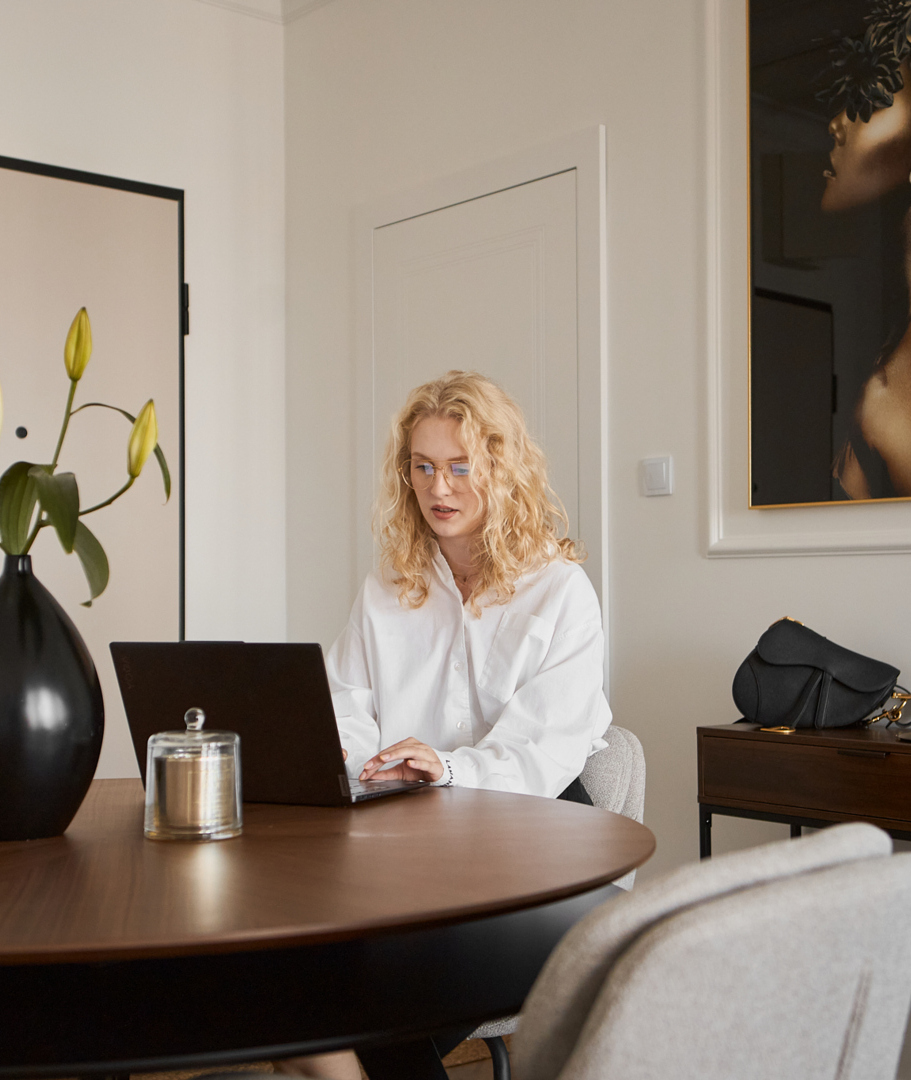 Gość pracujący na laptopie przy drewnianym stole w nowoczesnym apartamencie, czarny wazon z liliami, obraz i konsola z torebką.