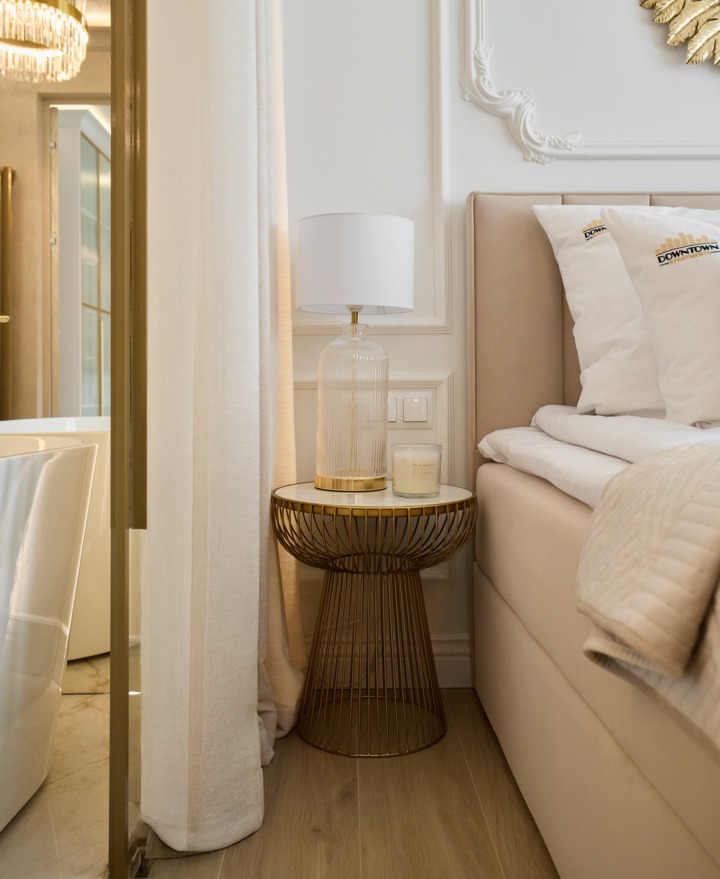 Tapicerowane łóżko z poduszkami z logo, złocisty stolik nocny z szklaną lampą i świecą, przyległa marmurowa łazienka