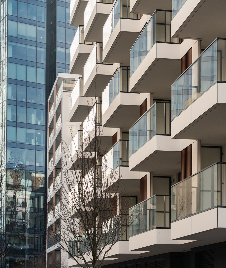 Nowoczesna fasada hotelu z przesuniętymi balkonami, szklanymi balustradami i drewnianymi akcentami obok szklanego wieżowca.