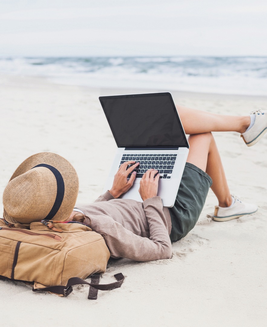 Persona sdraiata sulla spiaggia sabbiosa con laptop sulle gambe, cappello di paglia e zaino accanto, onde sullo sfondo