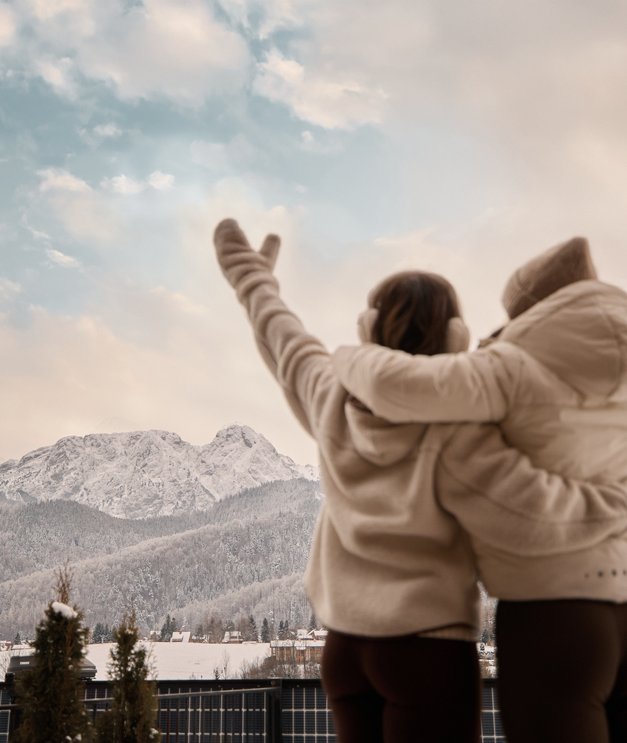 Dwoje gości na hotelowym balkonie, obejmują się z uniesionymi rękami, z widokiem na zaśnieżone góry i miasteczko.
