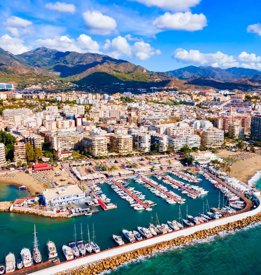 Porto turistico con yacht, spiagge di sabbia e hotel sul lungomare, montagne soleggiate e cielo azzurro.