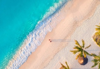 Pristine white sand beach with turquoise waves, scattered palm trees, and two people walking along the shoreline casting long shadows