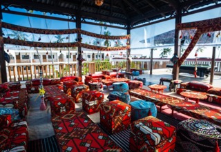 Open-air lounge with colorful patterned cushions, low tables, and hookahs under a wooden roof overlooking hotel balconies and palm trees