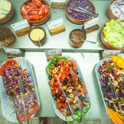 Colorful salad buffet with fresh vegetables, including cabbage slaw, tomato olive salad, lettuce, cucumbers, and various dressings in rustic bowls