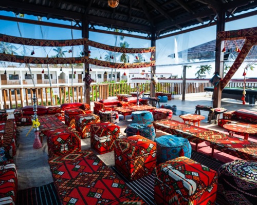 Open-air lounge with colorful patterned cushions, low tables, and hookahs under a wooden roof overlooking hotel balconies and palm trees