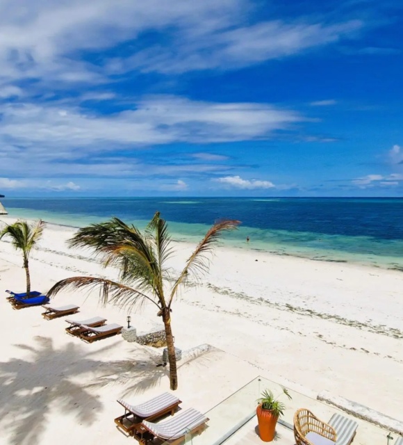 Tropical beachfront with white sand, palm trees, shaded daybeds, lounge chairs, and a sailboat on calm blue waters under a partly cloudy sky