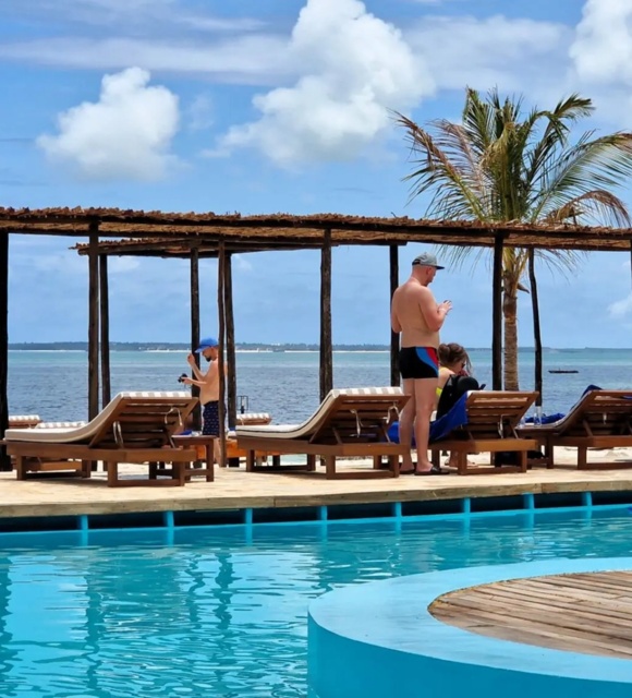 Oceanfront pool with curved blue edge, wooden lounge chairs, palm trees, and guests relaxing by the sea under a wooden pergola