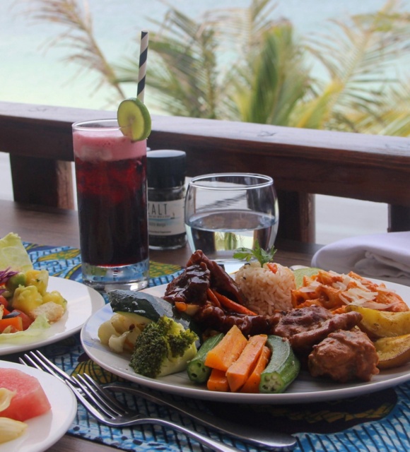 Tropical beachfront dining with plates of fresh salad, grilled vegetables, rice, and meat, plus a refreshing berry drink garnished with cucumber