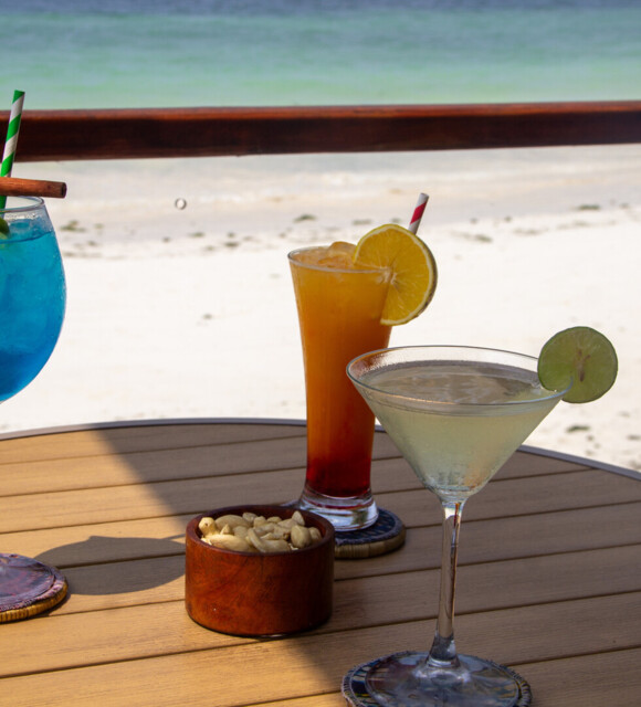 Three colorful cocktails with lime and lemon garnishes and a bowl of cashews on a wooden table overlooking a sandy beach and turquoise sea