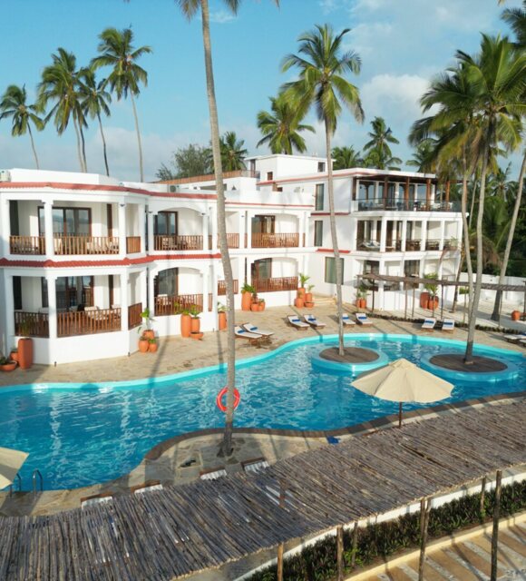 White three-story hotel with balconies overlooking a curved pool surrounded by palm trees and sun loungers under umbrellas