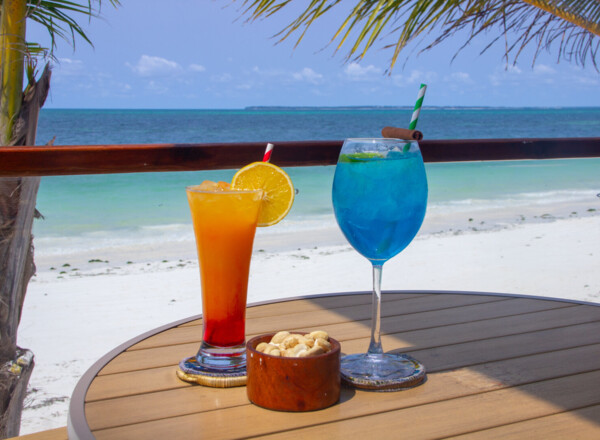 Tropical beachside table with vibrant orange and blue cocktails, a bowl of cashews, palm trees, and ocean view