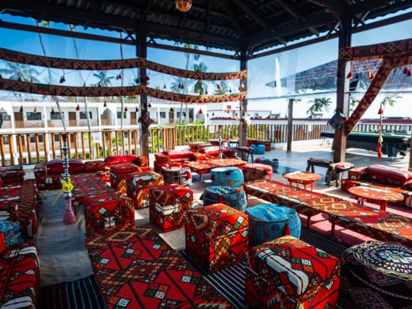 Open-air lounge with colorful patterned cushions, low tables, and hookahs under a wooden roof overlooking hotel balconies and palm trees