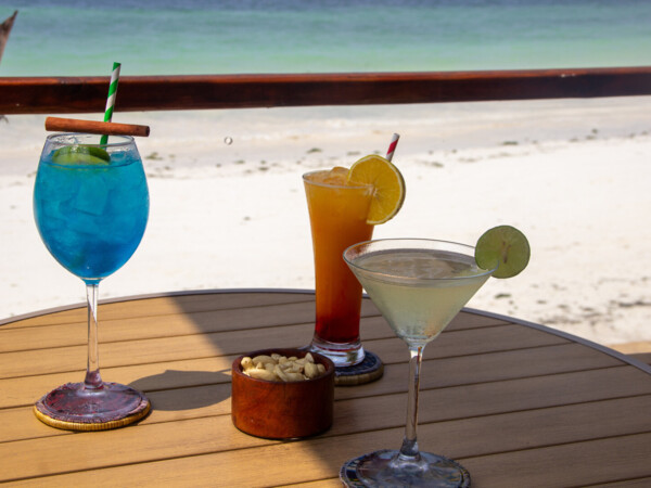 Three colorful cocktails with lime and lemon garnishes and a bowl of cashews on a wooden table overlooking a sandy beach and turquoise sea