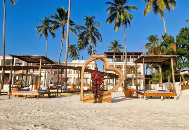 Beachfront resort with white sandy shore, palm trees, wooden shaded lounges, and a guest posing by a large heart-shaped frame