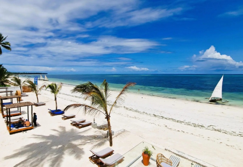 Tropical beachfront with white sand, palm trees, shaded daybeds, lounge chairs, and a sailboat on calm blue waters under a partly cloudy sky