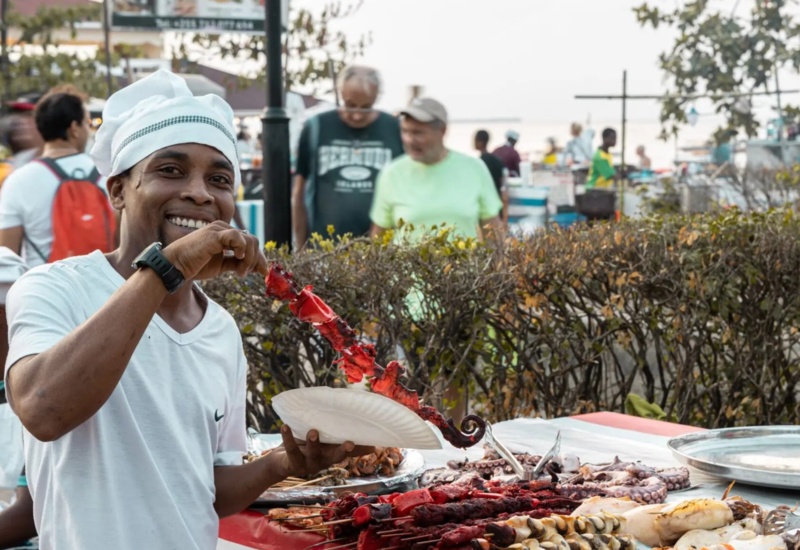 Street vendor with chef hat serving grilled octopus skewers at an outdoor food market near a leafy hedge