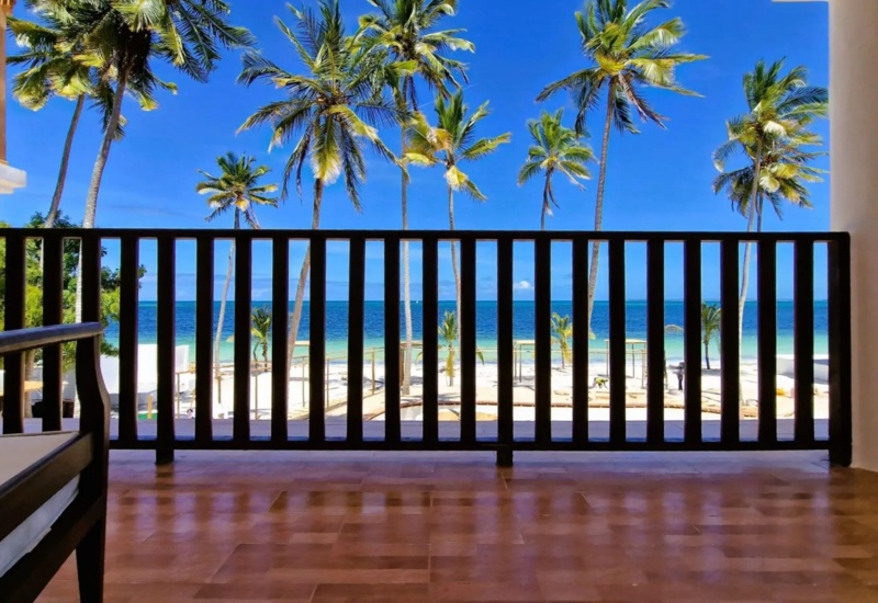Spacious balcony with wooden railing overlooking palm trees, white sandy beach, and clear blue ocean under a bright sky