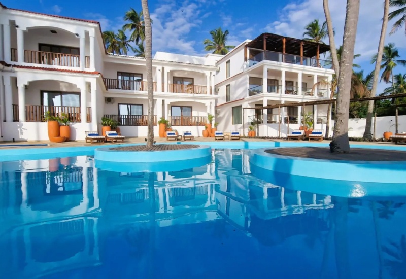 Resort with white Mediterranean-style buildings surrounding a large blue pool with palm trees and sun loungers on the deck