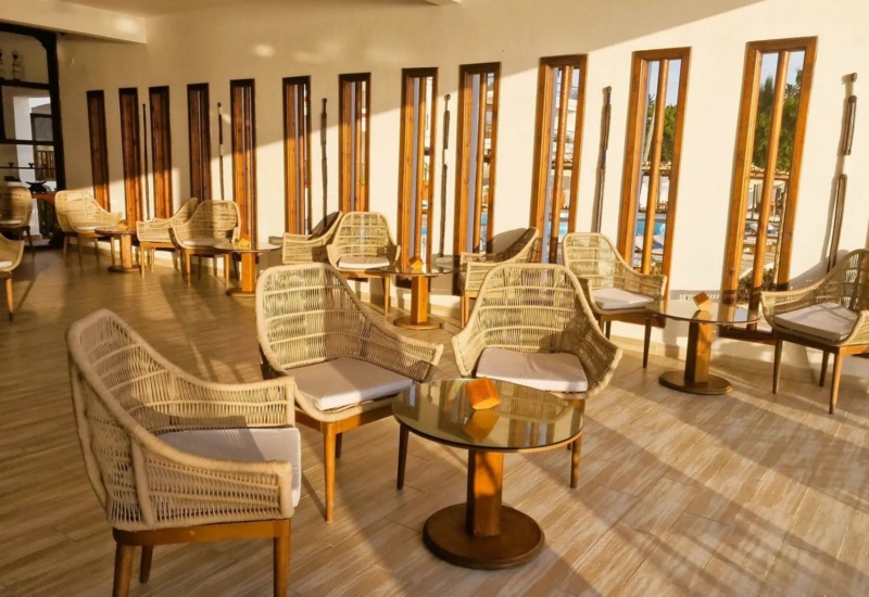 Sunlit hotel lounge with wicker chairs, glass-top tables, tall narrow windows, and warm wooden accents creating a relaxing atmosphere