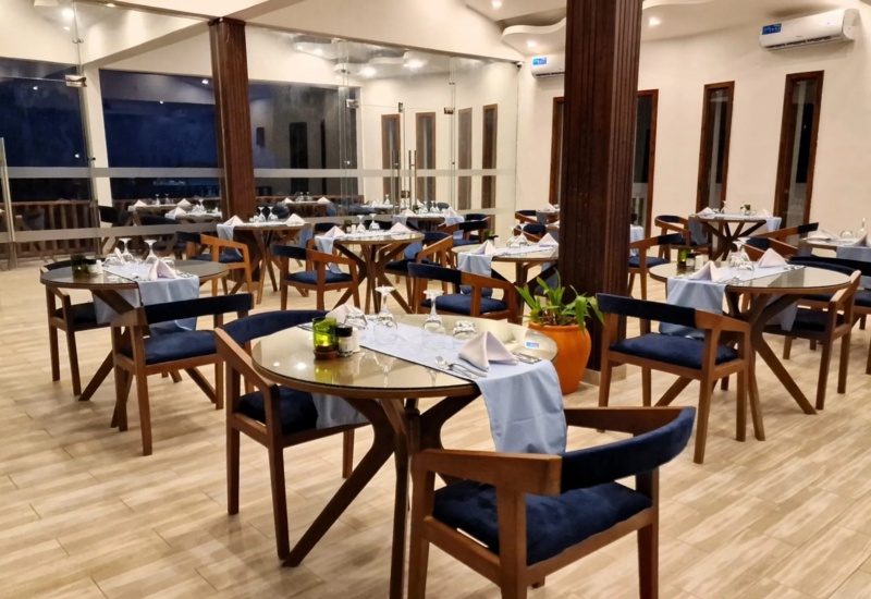 Elegant hotel dining room with wooden tables and chairs, blue cushions, white table runners, glassware, and modern decor