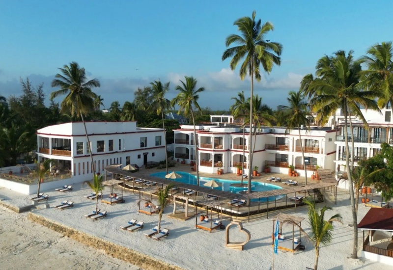 Beachfront resort with white modern buildings, central pool, tall palm trees, lounge beds under shaded pergola on sandy shore