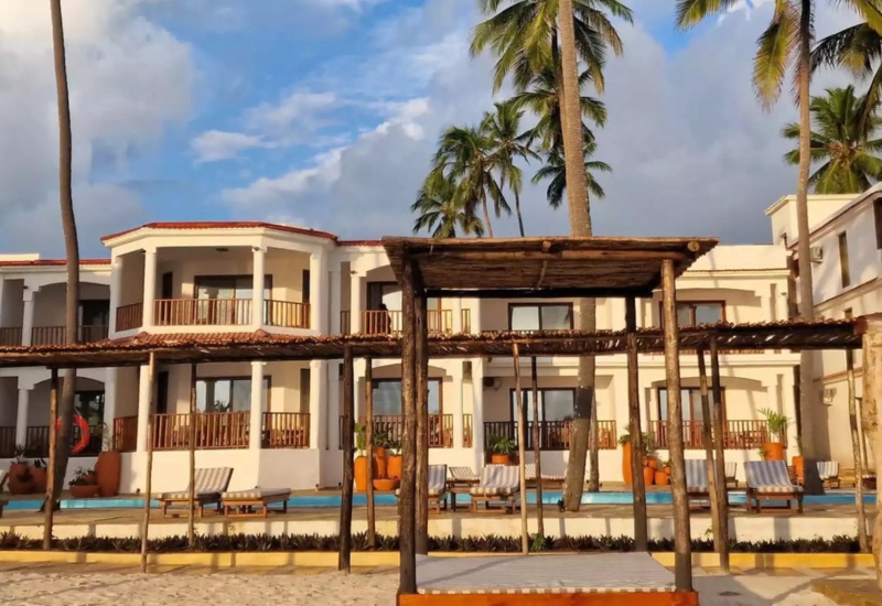 Beachfront hotel with white balconies, palm trees, wooden cabanas, and blue pool surrounded by lounge chairs under clear skies