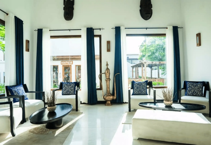 Bright hotel lounge with white armchairs, navy blue patterned cushions, large windows, and a decorative brass pitcher centerpiece