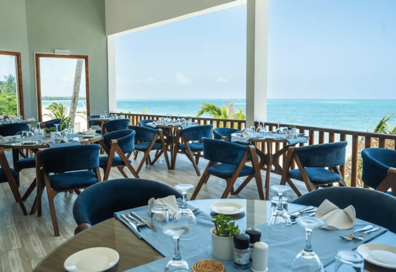 Seaside restaurant terrace with blue cushioned chairs, wooden tables set for dining, offering ocean views and tropical greenery