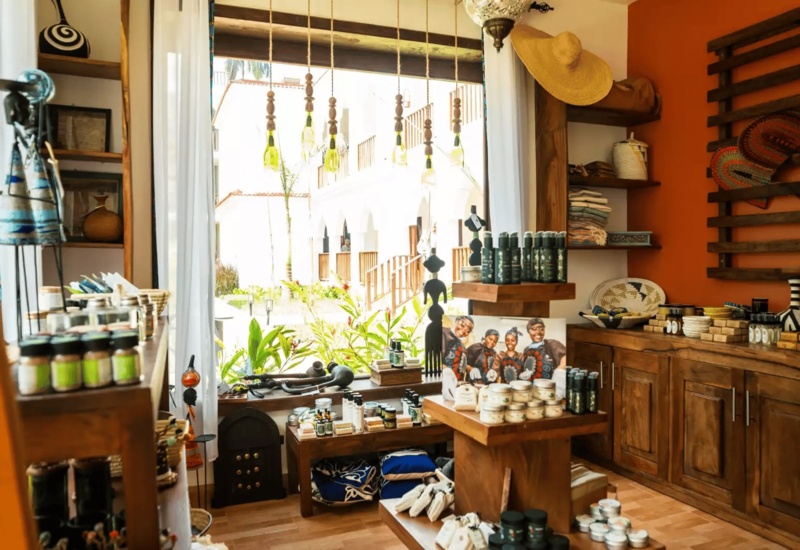 Boutique hotel gift shop with wooden shelves displaying local crafts, skincare products, textiles, and decorative items near a sunlit window.