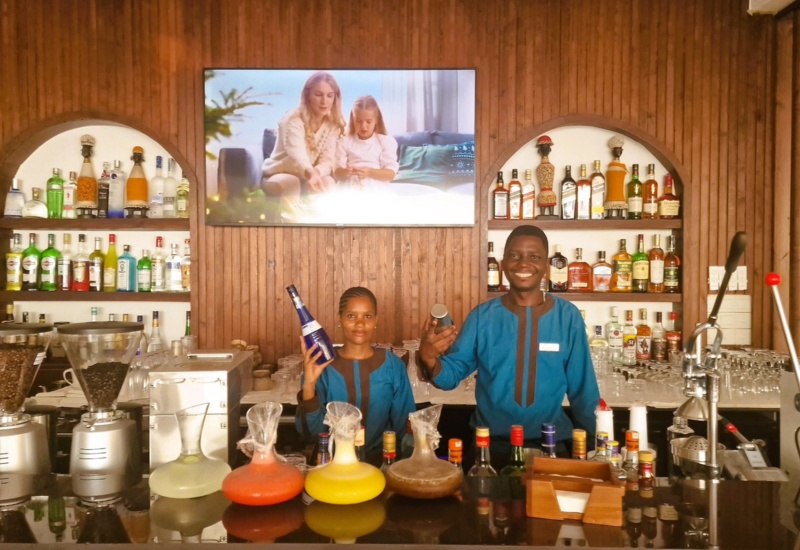 Two bartenders in blue uniforms smile behind a wooden bar stocked with colorful cocktails, liquor bottles, and coffee machines
