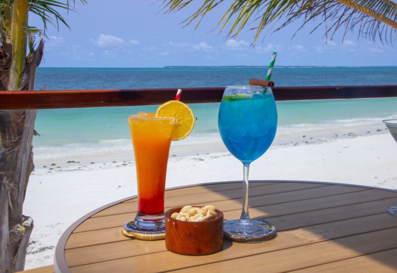 Tropical beachside table with vibrant orange and blue cocktails, a bowl of cashews, palm trees, and ocean view