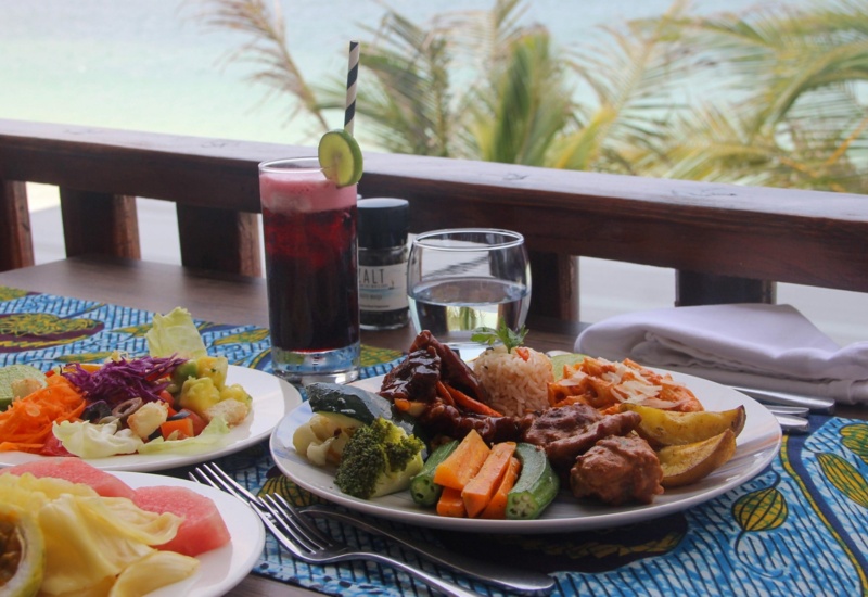 Tropical beachfront dining with plates of fresh salad, grilled vegetables, rice, and meat, plus a refreshing berry drink garnished with cucumber
