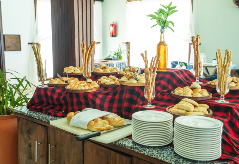 Breakfast buffet with assorted fresh breads, breadsticks in tall glasses, white plates stacked neatly on a red plaid-covered table