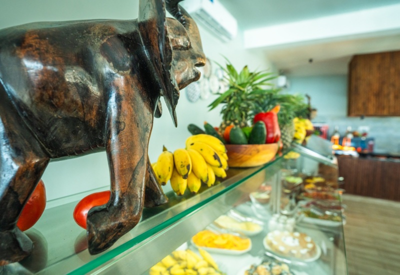 Wooden elephant sculpture and fresh tropical fruit display, including bananas and peppers, in a hotel breakfast buffet area