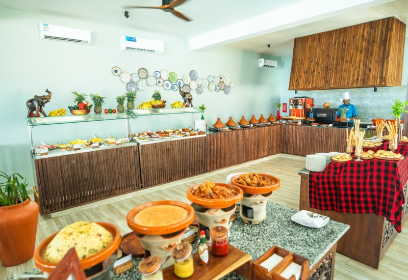 Buffet dining area with traditional dishes in clay pots, fresh fruit displays, warm wooden decor, and a chef preparing meals