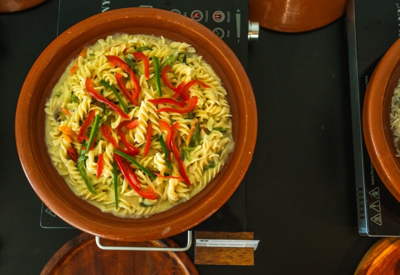 Twisted pasta in creamy sauce with sliced red and green peppers served in a rustic clay bowl on a buffet station