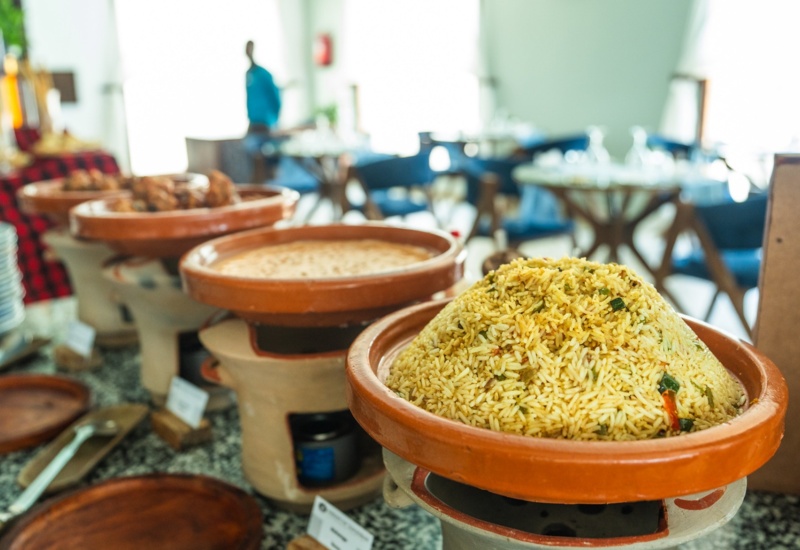 Buffet setup with large clay pots holding seasoned rice and other hot dishes in a bright dining area with blue chairs and wooden tables