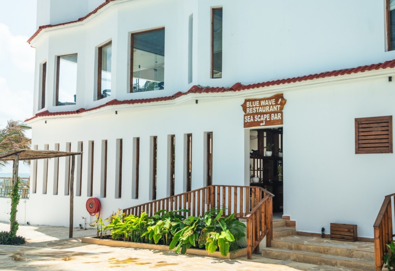 White beachfront hotel facade with wooden railings, tropical plants, and signs directing to Blue Wave Restaurant and Sea Scape Bar