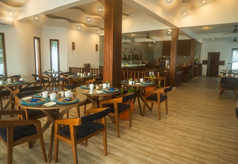 Bright hotel restaurant with wooden tables and chairs, set for breakfast with cups, plates, and blue napkins, featuring warm lighting