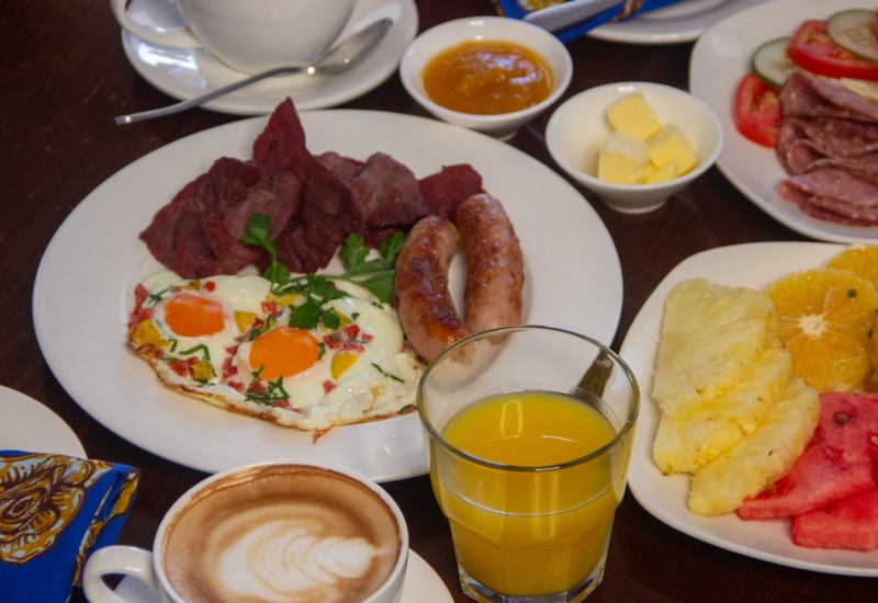 Hearty breakfast with fried eggs, sausage, bacon, fresh fruit platter, orange juice, coffee, and butter on a wooden table