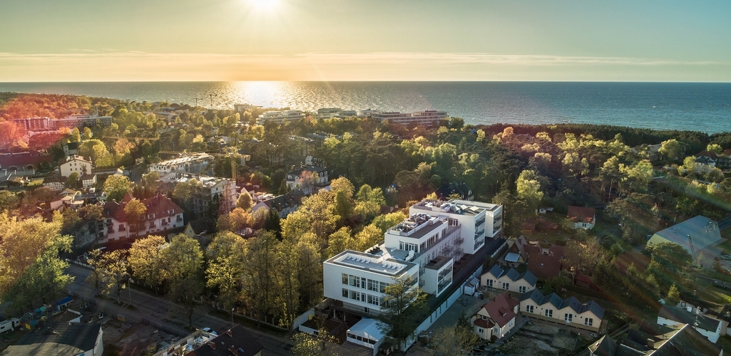 Modern white seaside hotel with rooftop terraces overlooking the sunlit sea, set among tree-lined villas and coastal forest.