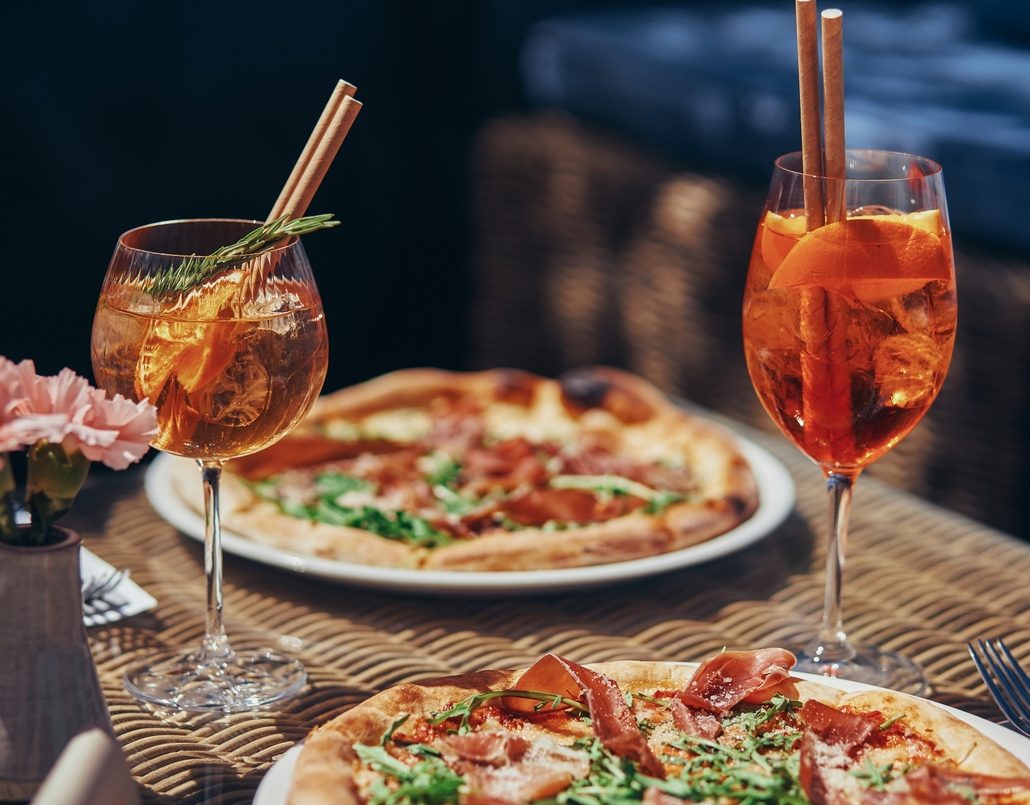 Two Aperol-style spritzes with orange and rosemary beside wood-fired pizzas on a wicker table, al fresco terrace dining.