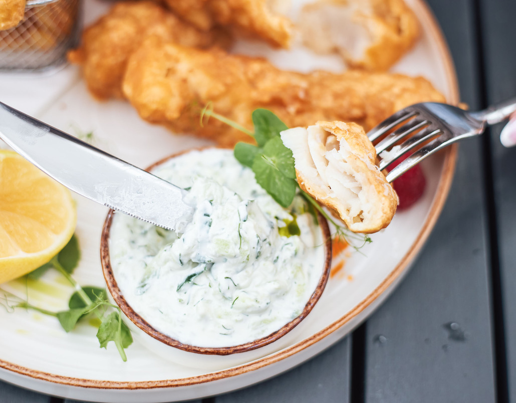 Hand lifting a fork with a bite of crispy battered fish, lemon wedge, creamy herb tartar and fries on a plated seafood dish