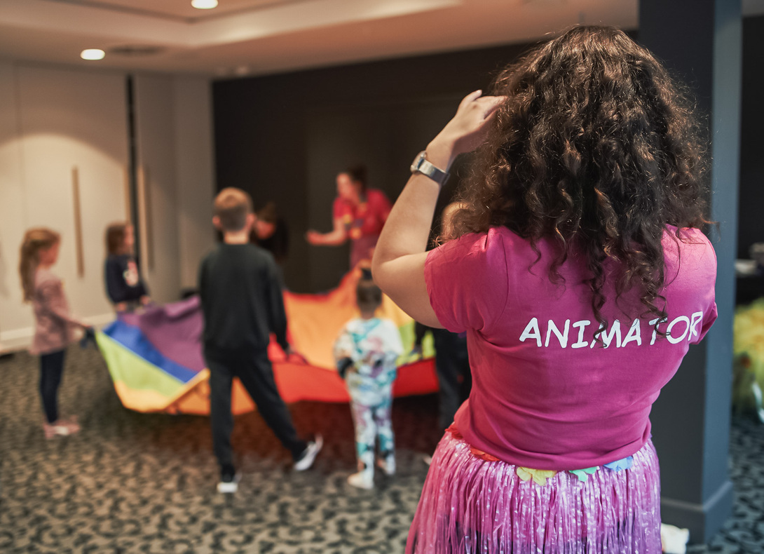 Animatorka w różowej koszulce i spódnicy hula prowadzi dzieci w zabawie z kolorowym spadochronem w sali aktywności