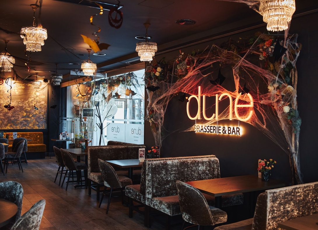 Cozy brasserie interior with velvet booths, chandeliers and neon 'dune Brasserie & Bar' sign, Halloween cobwebs and bat decor