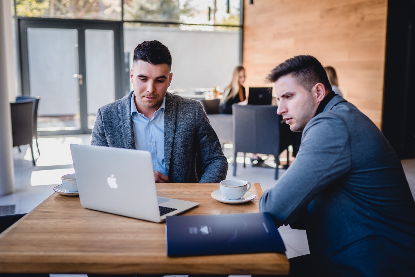 Two men holding a business meeting at a hotel lounge table with a laptop, coffee cups and bright modern wood interior.