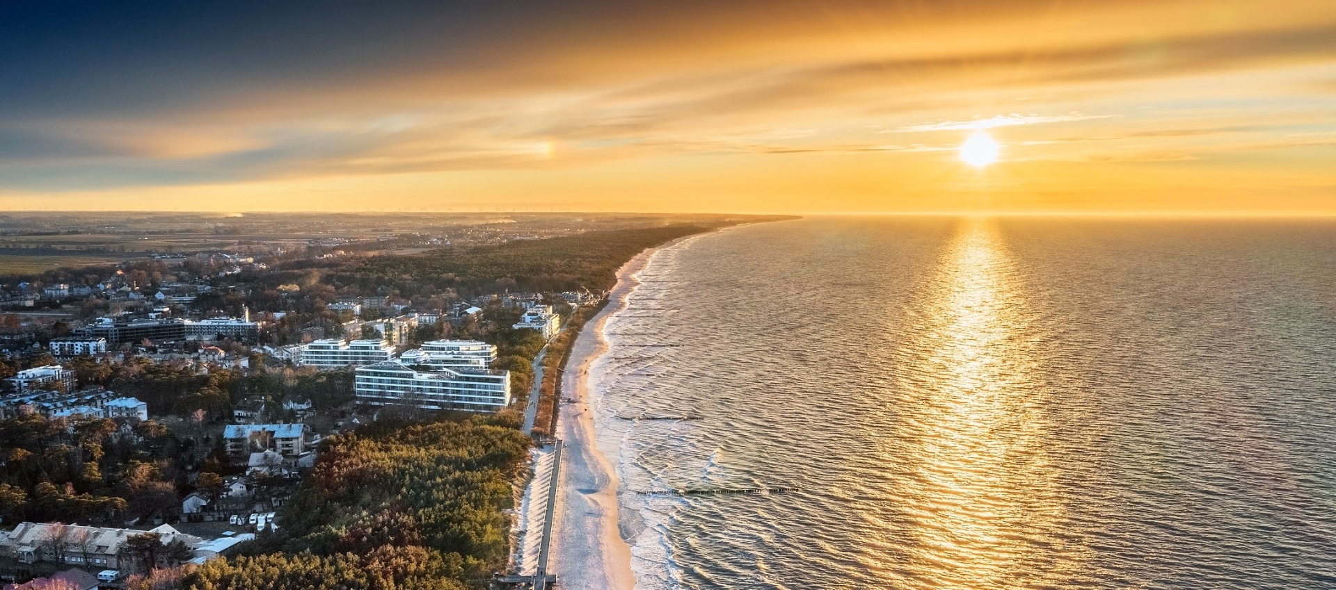 Aerial view of beachfront hotels and promenade along a long sandy coast at golden sunset, calm sea reflecting warm light.