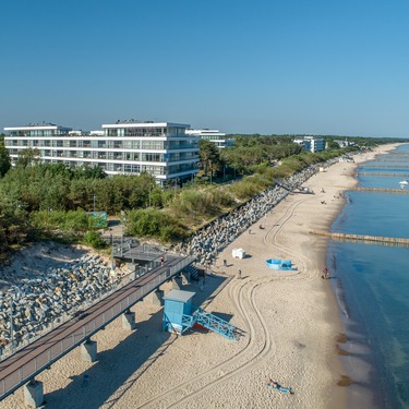 Nowoczesny hotel przy plaży obok sosnowego lasu, z promenadą, piaszczystą plażą, wieżą ratownika i falochronami.